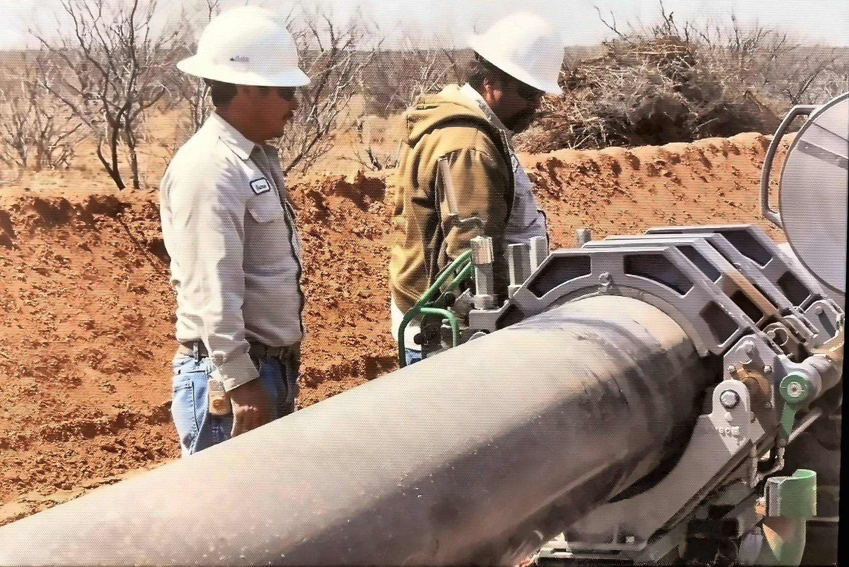Two Encore crew members in hard hats beside a steel pipeline tie-in